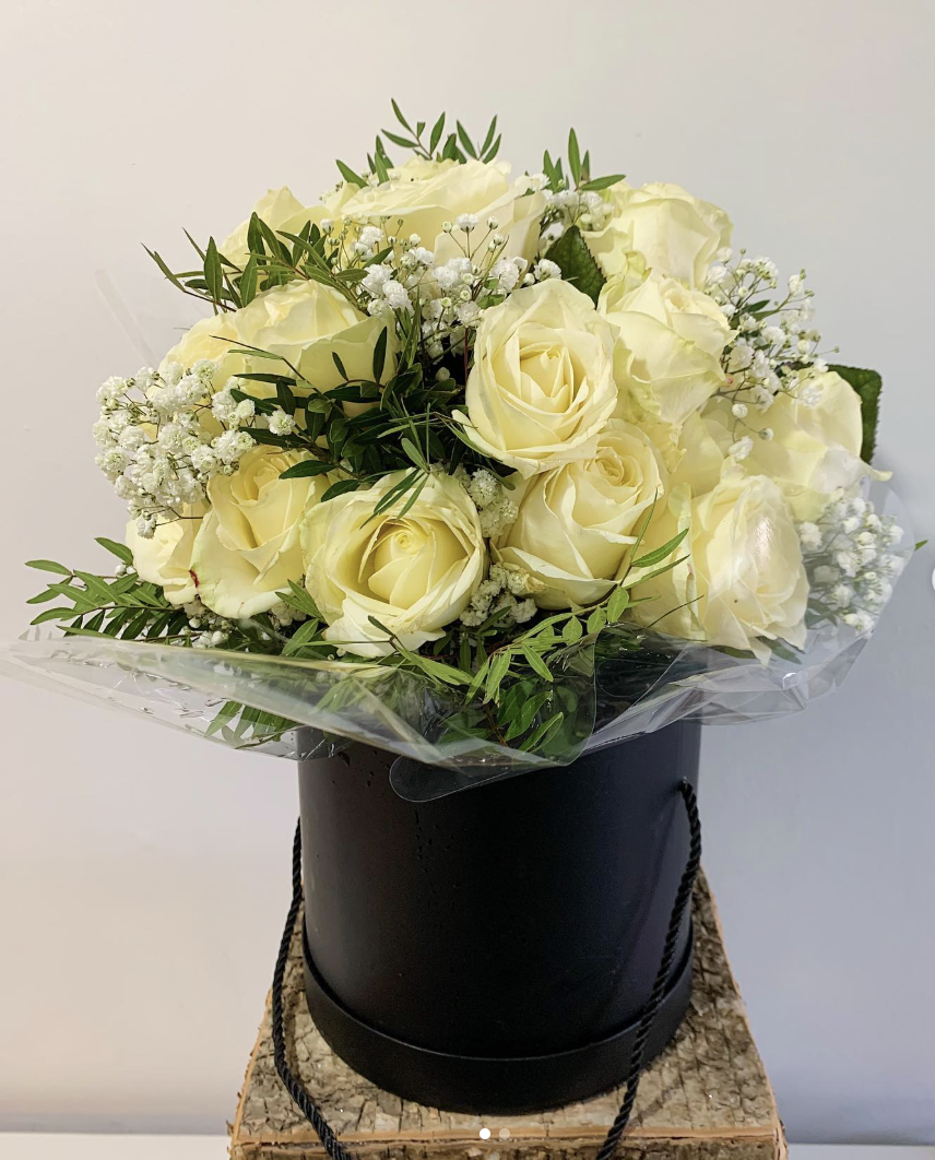 A hand tied bouquet of ivory roses and white gypsophila arranged in a round black hat box, finished with trailing foliage