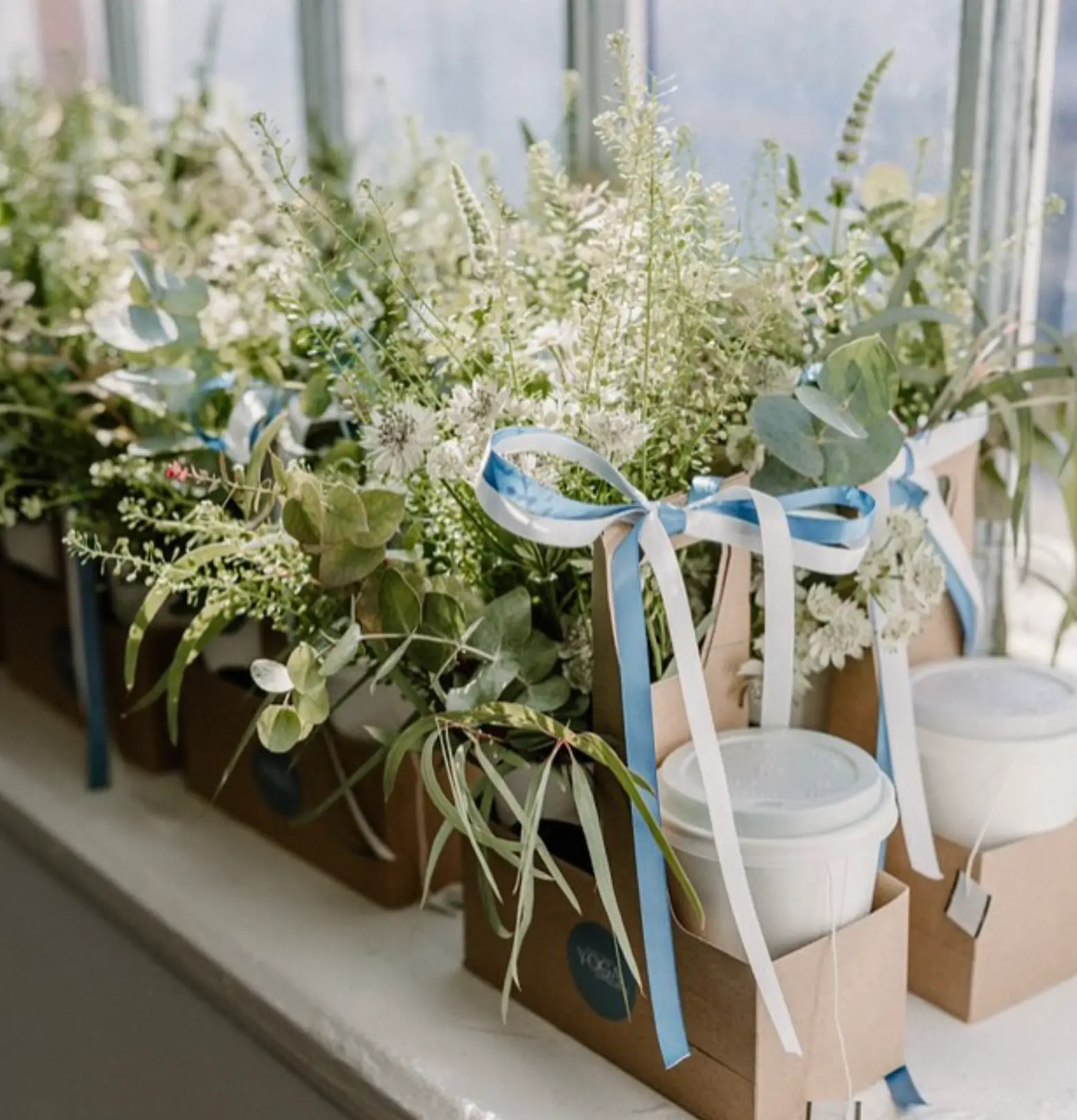 Corporate gift bags lined up on a windowsill, each containing fresh white and green flowers tied with blue and white ribbons alongside branded takeaway coffee cups