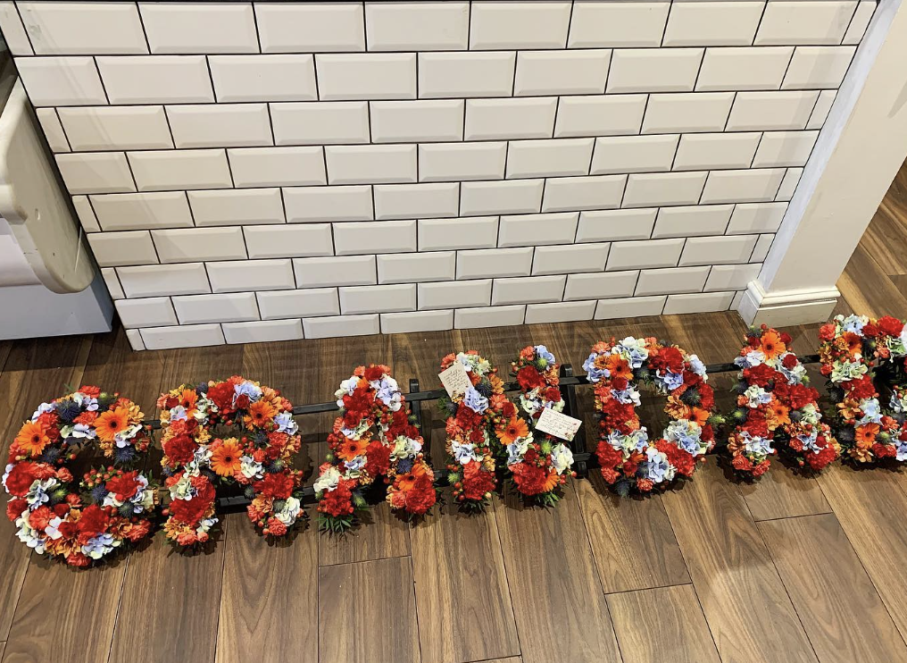 A funeral tribute made of fresh wreaths spelling GRANDAD, in reds, oranges, white and blue flowers, arranged on a wooden floor