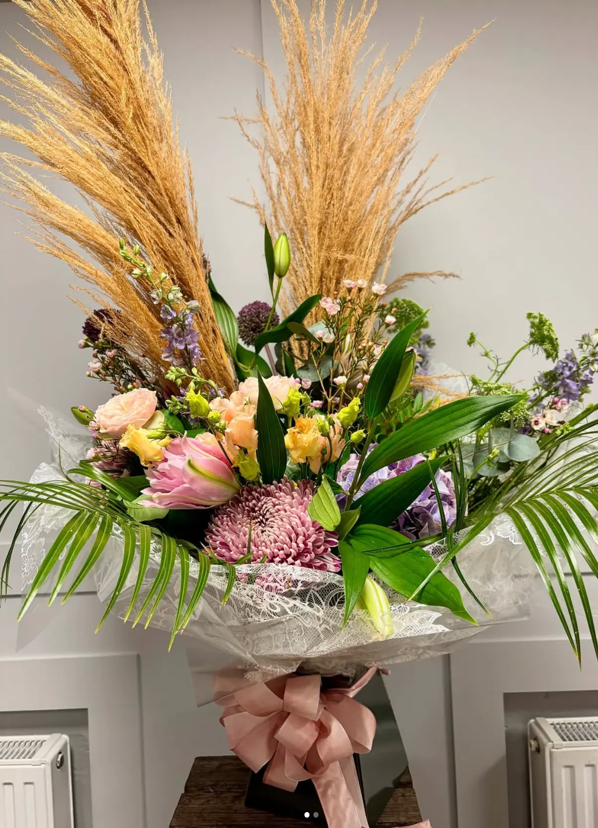 A celebratory hand tied bouquet of pampas grass, pink and peach roses, purple alliums, hydrangea and trailing foliage, wrapped in lace and tied with a pink satin bow