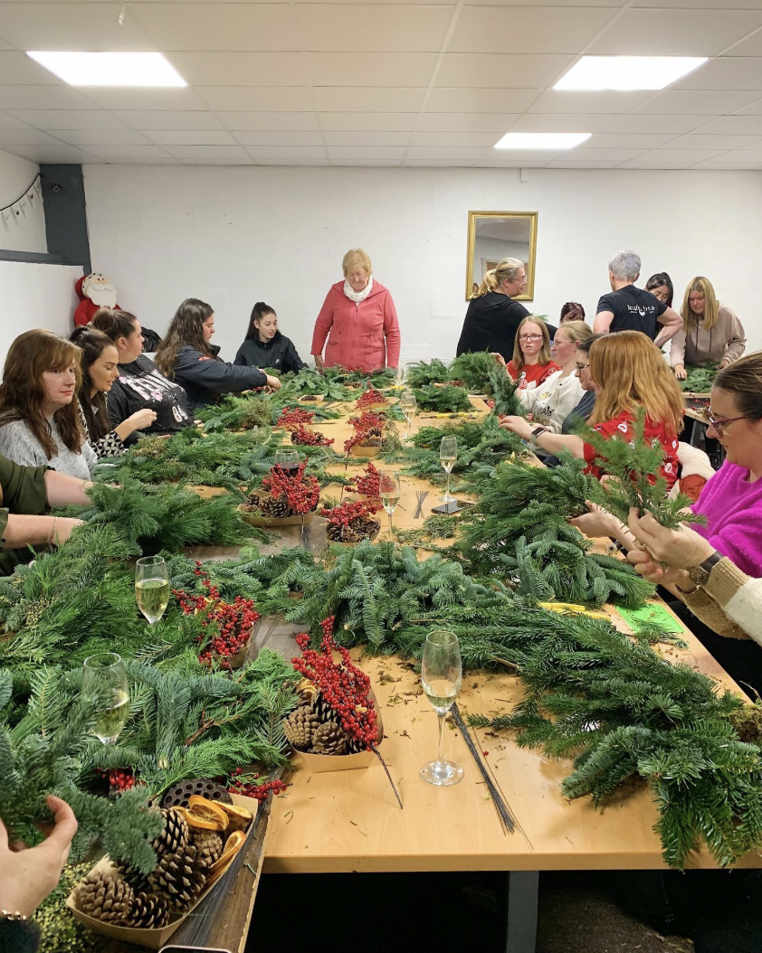 A group of people making Christmas wreaths at a Leaf + Bean Florist workshop, surrounded by fresh greenery, berries and pine cones on a long table