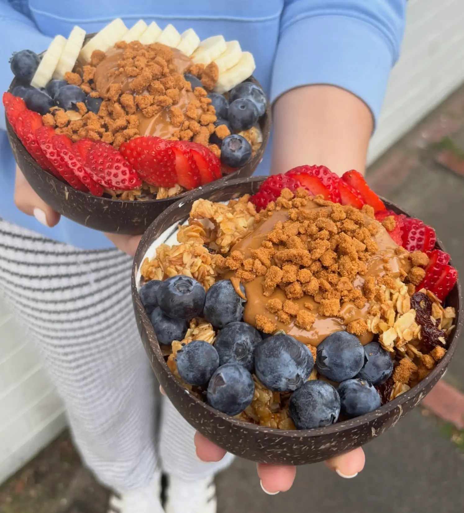 Two coconut bowls of granola, berries, banana and Biscoff crumb held up outside the cafe