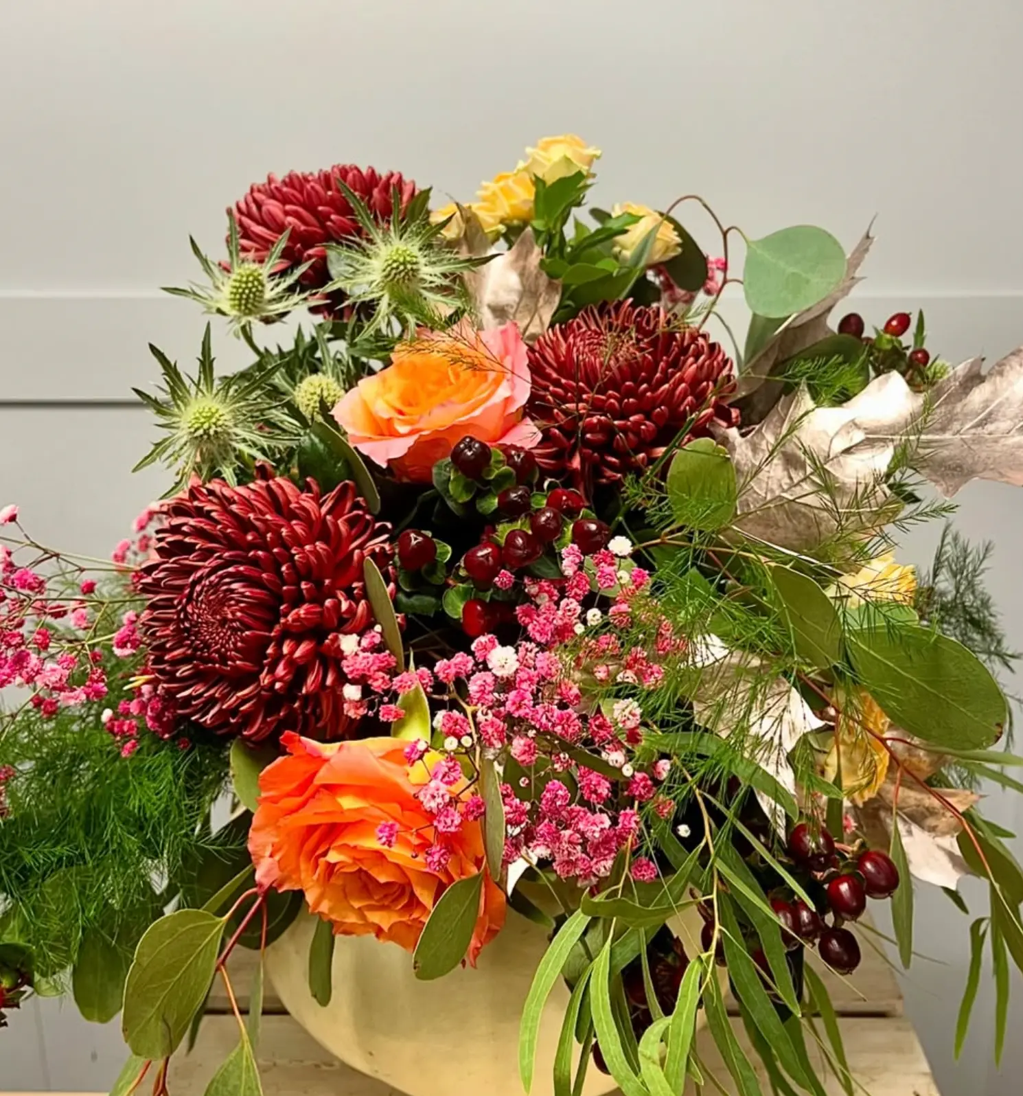 A burgundy and orange arrangement of chrysanthemums, roses, thistle and hypericum berries