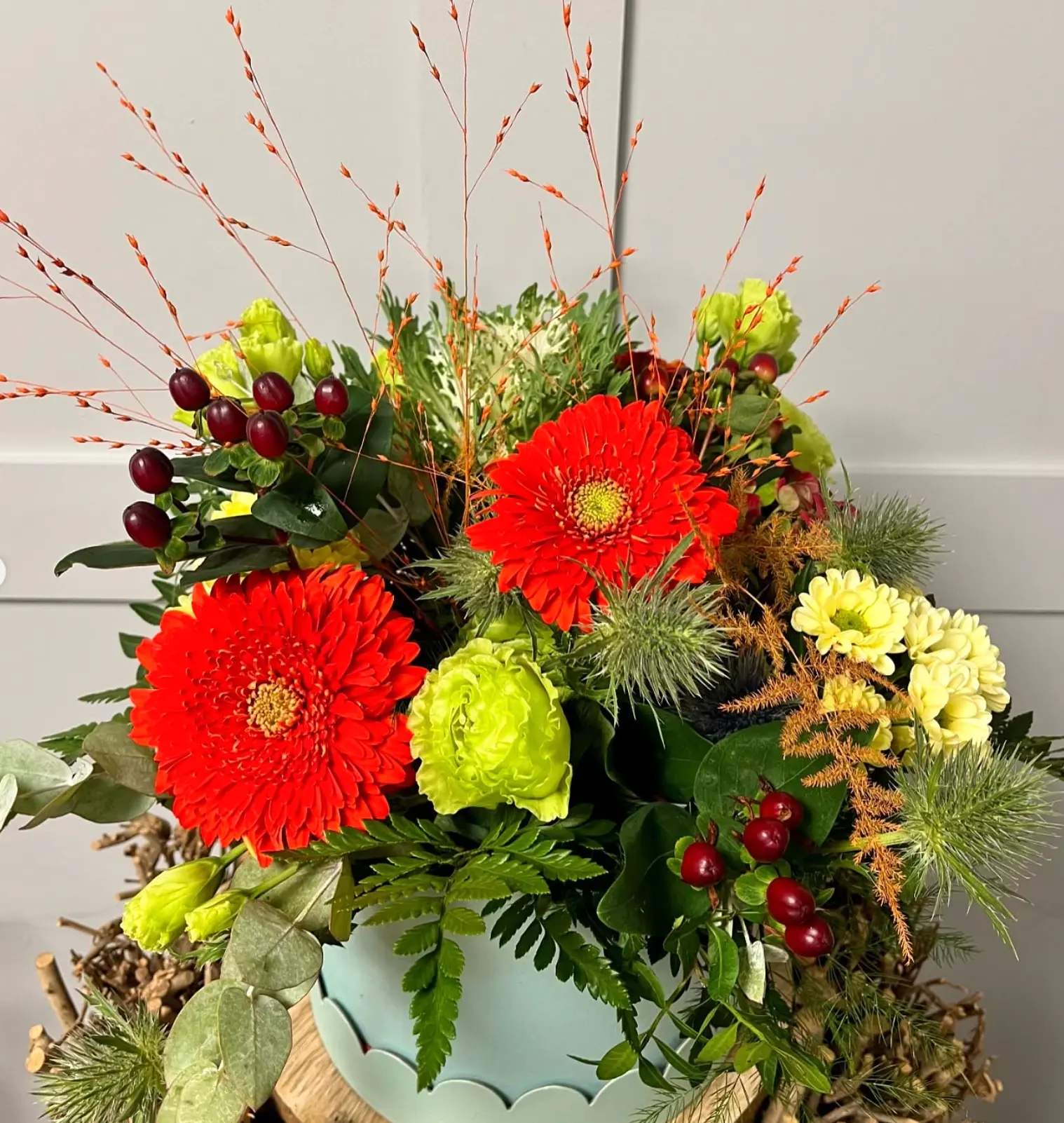 A pale blue scalloped pot filled with red gerberas, green carnations, thistle and hypericum berries