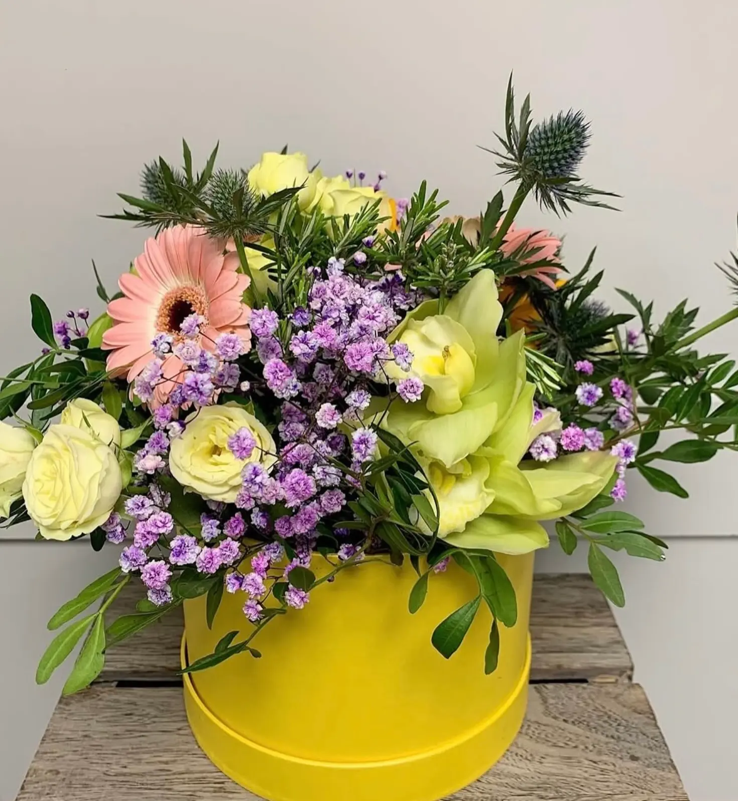 A yellow hat box arrangement of cream roses, pink gerbera, orchid and purple statice