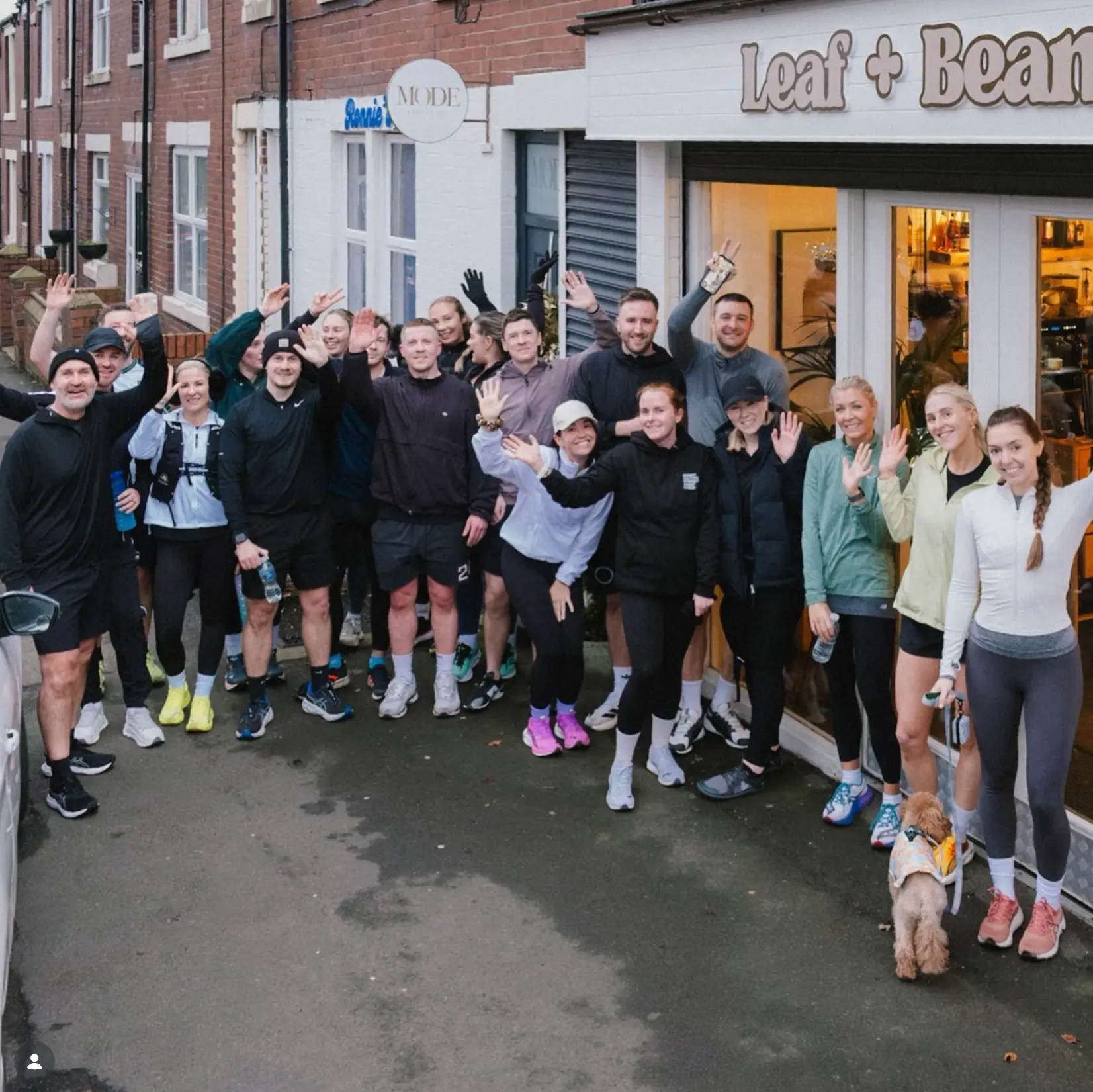 A large group of run club runners waving outside the Leaf + Bean shopfront on Emmerson Terrace, with a small dog on a lead beside them